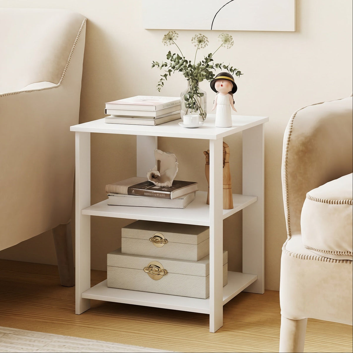A small white three-tier side table styled in a cozy living room setting, flanked by two beige armchairs. The table is decorated with books, a small vase of greenery, a doll, and storage boxes.